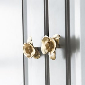 May include: Two gold-toned flower-shaped cabinet knobs. The knobs are made of metal and have a realistic flower design.