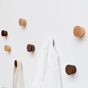 Puede incluir: Un conjunto de ganchos de pared de madera en varios tonos de marrón y beige. Los ganchos son de forma cilíndrica y están montados en una pared blanca. Una prenda blanca está colgada en uno de los ganchos.