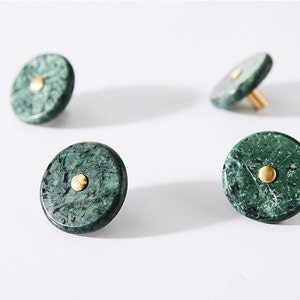 Op de afbeelding: Vier groene marmeren kastknoppen met messing accenten. De knoppen zijn rond met een platte voorkant en een kleine messing stip in het midden. Het marmer heeft een donkergroene kleur met lichtere groene adering. Voor meubels.