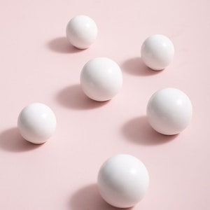 Könnte beinhalten: Sechs weiße Keramikkugeln, die in einem versetzten Muster auf einem hellrosa Hintergrund angeordnet sind.