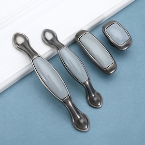 Può includere: Set di quattro pomelli per armadi in ceramica grigia con finitura argentata. I pomelli sono di forme e dimensioni diverse, tra cui due tiretti ovali lunghi e due pomelli quadrati.