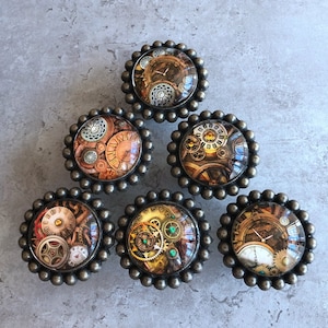 May include: Six steampunk cabinet knobs with clock face designs. Each knob has a domed glass centre with gears and clock faces in brown, gold, and white, set in a bronze-coloured metal base with a beaded edge.