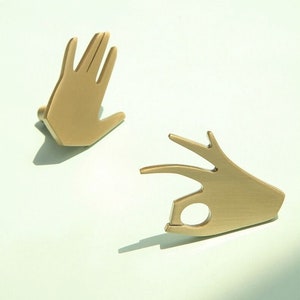 May include: Two gold-coloured metal hand-shaped drawer pulls. One pull is shaped like a hand with all fingers extended, the other is shaped like a hand making the 'okay' sign.