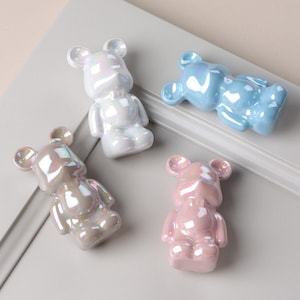 May include: Four ceramic bear drawer pulls in different colours: white, light blue, light pink, and grey. Each bear has a glossy finish and iridescent details.