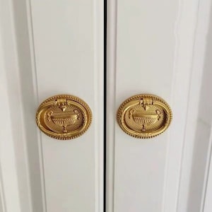 Può includere: Due pomelli per porte ovali dorati con un design decorativo che presenta un'urna stilizzata con un motivo floreale.