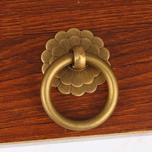 Può includere: Un tirarello ad anello in ottone con un design floreale decorativo. Il tirarello ad anello è attaccato a una superficie in legno.