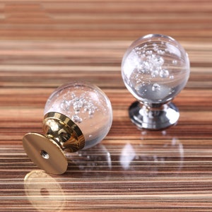 以下が含まれることがあります： 金色のアクセントと銀色のベースが付いた、透明なガラス製のキャビネットノブが2つ。ガラスには小さな気泡が入っています。