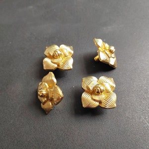 Op de afbeelding: Vier gouden metalen knoppen in de vorm van een bloem. Elke knop heeft een verhoogd midden en een gestructureerd ontwerp.
