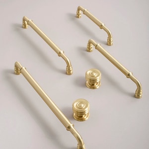 Puede incluir: Un conjunto de cinco piezas de herrajes para armarios de color dorado, que incluyen cuatro tiradores y un pomo. Los tiradores son de diferentes longitudes y tienen un diseño redondeado ligeramente curvado. El pomo es redondo y tiene una superficie texturizada.