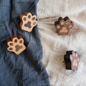 May include: Four wooden paw print hooks in brown and light brown. The hooks are in the shape of a paw print and have a small metal loop on the back for hanging.