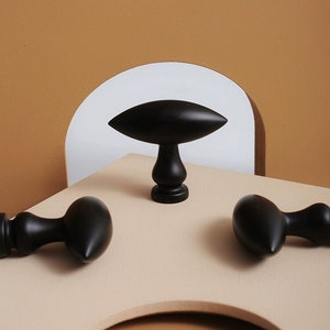 May include: Three black wooden cabinet knobs with a unique teardrop shape. The knobs are on a beige surface with a brown background.