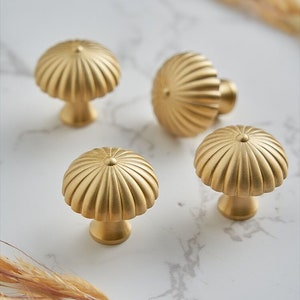 Op de afbeelding: Vier gouden kastknoppen met een geribbeld, gegolfd ontwerp. De knoppen zijn gerangschikt op een wit marmeren oppervlak.