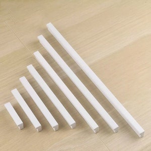 May include: Set of seven white cabinet pulls in various sizes. The pulls are rectangular with a square end and a flat surface.