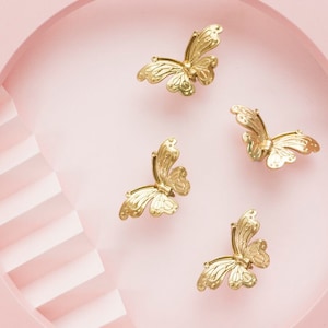 Könnte beinhalten: Vier goldene Schmetterlingshaarklammern auf rosa Hintergrund. Die Schmetterlinge sind in einem Kreis angeordnet, wobei die Flügel jedes Schmetterlings nach außen zeigen.