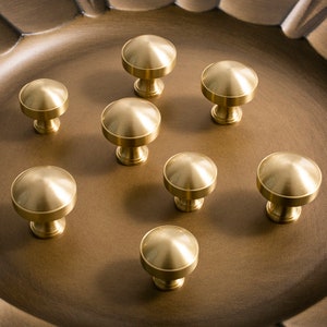 以下が含まれることがあります： 丸みを帯びたドーム型のトップと円筒形のベースを持つ、金色のキャビネットノブが8つ。ノブは茶色の表面に配置されています。