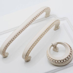 Op de afbeelding: Drie witte kastknoppen met gouden rand. De knoppen zijn in de vorm van een ring, een lange handgreep en een kortere handgreep.