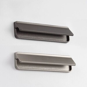 Może przedstawiać: Dwa prostokątne uchwyty do szafek z wykończeniem w szczotkowanym niklu. Uchwyty mają wpuszczany uchwyt i lekko pochylony brzeg.