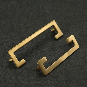Peut inclure: Deux poignées de meuble en métal doré avec une forme rectangulaire et une découpe carrée au centre. Les poignées ont une finition brossée.
