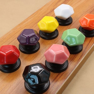 以下が含まれることがあります： 幾何学的なダイヤモンドの形をした、カラフルなセラミック製のキャビネットノブのセット。各ノブは黒いベースに取り付けられています。