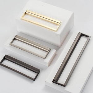 May include: Four cabinet pulls in different finishes: gold, nickel, and two shades of grey. The pulls are rectangular with a recessed centre.