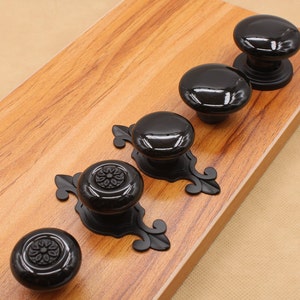 Może przedstawiać: Pięć czarnych gałek do szafek z ozdobnymi podstawami metalowymi. Gałki są okrągłe i mają błyszczące wykończenie. Dwie z gałek mają ozdobny wzór kwiatowy.