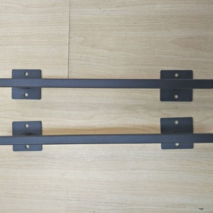 May include: Two black metal door handles with square ends. The handles are mounted on a light wood surface.