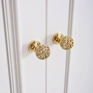 以下が含まれることがあります： 装飾的な透かし彫りのデザインが施された、金色のキャビネットノブが2つ。ノブは丸型で、滑らかで磨かれた仕上げになっています。