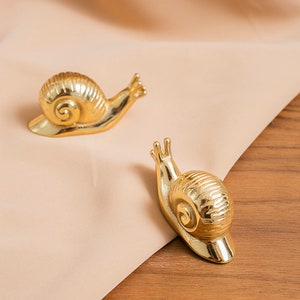 Può includere: Due figurine di lumaca dorate su uno sfondo di tessuto marrone chiaro. Le lumache sono rivolte in direzioni diverse e hanno un guscio testurizzato e dettagliato.