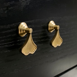 Op de afbeelding: Twee gouden kastgrepen met een ginkgo-bladontwerp. De grepen zijn gemonteerd op een zwart oppervlak.