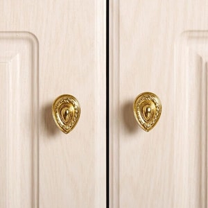 Peut inclure: Deux boutons de porte en or décoratifs avec un design orné. Les boutons sont en forme de larme avec un centre surélevé.