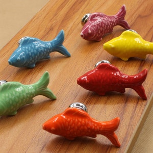 May include: Six ceramic fish drawer pulls in various colours, including blue, green, yellow, red, and orange. Each pull has a silver metal knob and a realistic fish design.