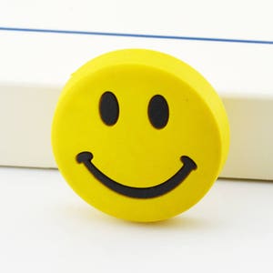 Pode incluir: Uma borracha amarela em forma de carinha sorridente com contorno preto. A borracha é redonda e tem uma expressão feliz.