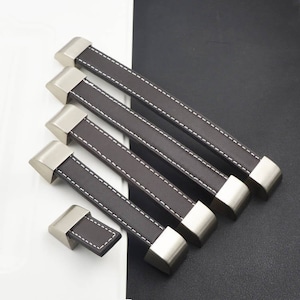 Peut inclure: Ensemble de poignées de meubles en cuir marron avec des coutures blanches et des embouts en nickel brossé. Les poignées varient en taille, la plus longue mesurant environ 20 cm de long. L'ensemble est disposé en diagonale.