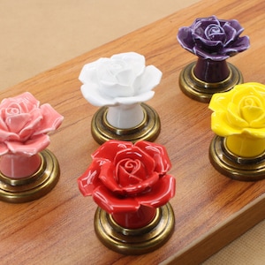 May include: Five ceramic rose-shaped drawer pulls with gold-tone bases. The roses are in shades of pink, white, yellow, red, and purple.