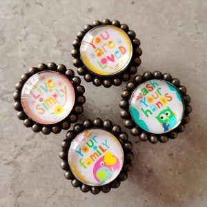 May include: Four round, bronze-coloured cabinet knobs with glass inserts. Each knob features a different colourful design with a positive message. The messages are "You are loved", "Live Simply", "Wash Your Hands", and "Love Your Family".