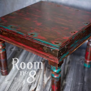 Könnte beinhalten: Ein kleiner, quadratischer Holztisch mit einer abgenutzten roten, braunen und türkisfarbenen Lackierung. Der Tisch hat vier Beine mit gedrehtem Design und Metallverzierungen am oberen Rand.