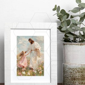 May include: A framed print depicts a figure in white robes with a child in a pink dress, set in a field of flowers. The artwork has a soft, painterly style, with a white wooden frame. A potted plant is visible on the right.