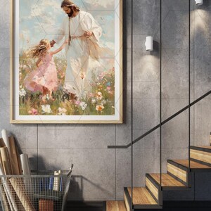May include: A framed art print depicts a figure in white robes with a child in a pink dress in a field of flowers. The artwork is in a light wood frame, displayed on a gray tiled wall. The scene evokes a sense of peace and serenity.
