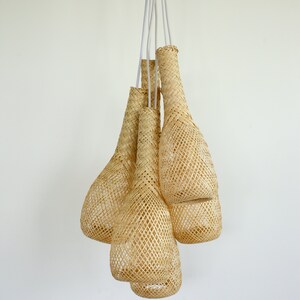 May include: A set of five woven pendant lights, each shaped like a teardrop, hanging from white cords against a white wall. The lights are made of natural woven material, giving them a rustic and organic look.