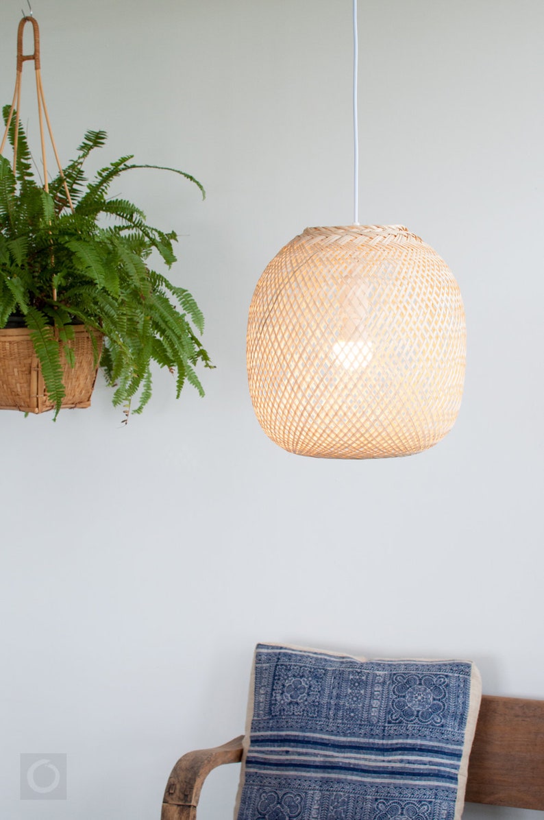 Op de afbeelding: Een geweven bamboe hanglamp hangt aan een wit snoer tegen een witte muur. De lamp is rond en heeft een lichtbruine kleur.