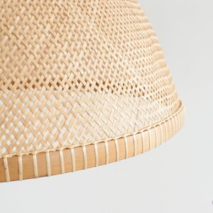 May include: Close-up of a natural woven lampshade with a light brown color. The lampshade is made of a woven material with a natural finish. The lampshade has a round shape and a slightly curved top.