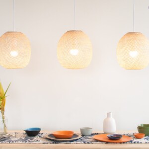 Op de afbeelding: Drie geweven hanglampen hangen boven een tafel die is gedekt met blauwe en witte placemats, oranje en blauwe kommen en een witte vaas met een groene en gele bloemschikking.