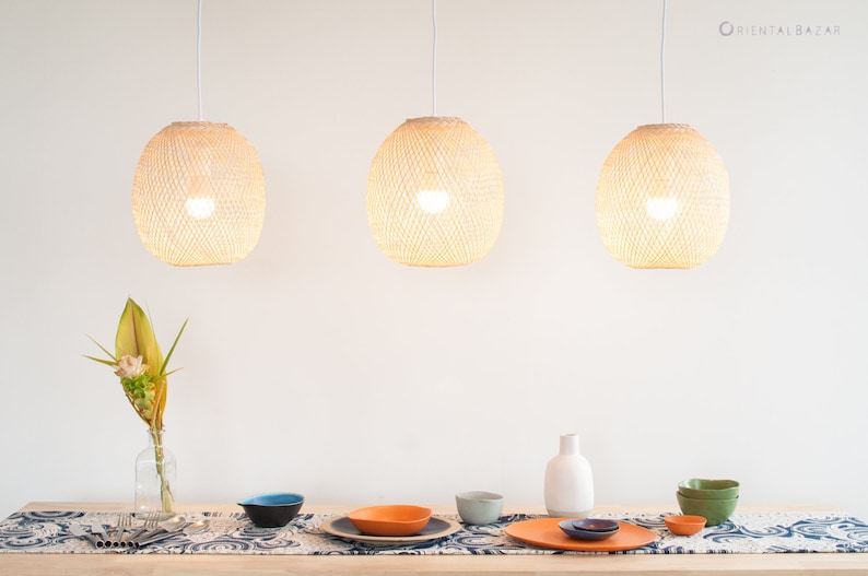 Op de afbeelding: Drie geweven hanglampen hangen boven een tafel die is gedekt met blauwe en witte placemats, oranje en blauwe kommen en een witte vaas met een groene en gele bloemschikking.