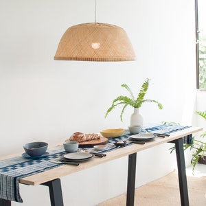 May include: A wooden dining table with a blue and white patterned table runner. The table is set with plates, bowls, and silverware. A woven lampshade hangs above the table. A potted plant is in the background.