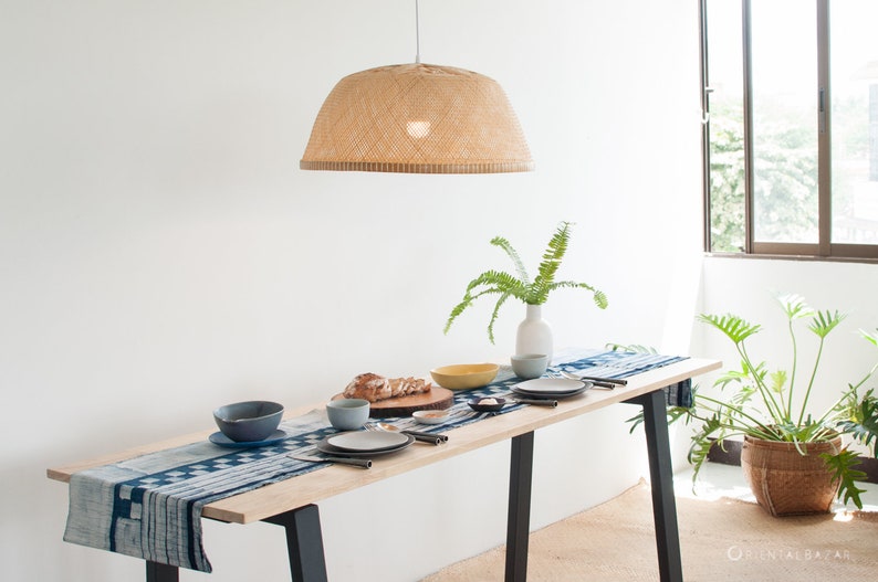 May include: A wooden dining table with a blue and white patterned table runner. The table is set with plates, bowls, and silverware. A woven lampshade hangs above the table. A potted plant is in the background.