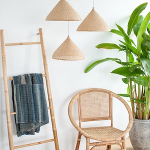 May include: Three woven pendant lights hang from the ceiling above a bamboo ladder with a blue and white throw blanket draped over it. A woven rattan chair sits in front of a potted green plant.