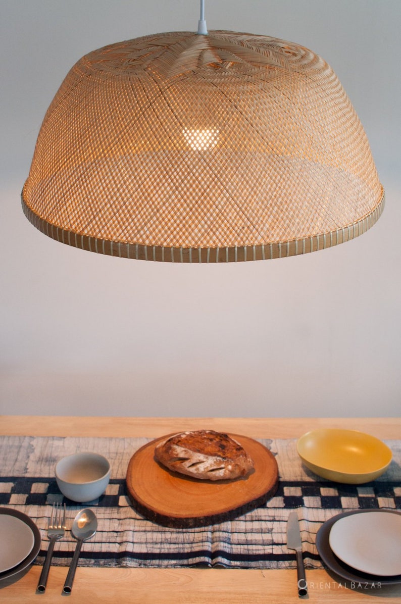 May include: A woven bamboo pendant light fixture hangs over a wooden table with a blue and white patterned table runner. A loaf of bread sits on a wooden cutting board on the table runner. There are two bowls and two plates on the table.