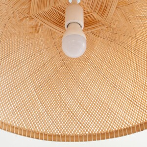 May include: A close-up of a woven wicker lampshade with a white light bulb inside. The lampshade is made of natural materials and has a light brown color.