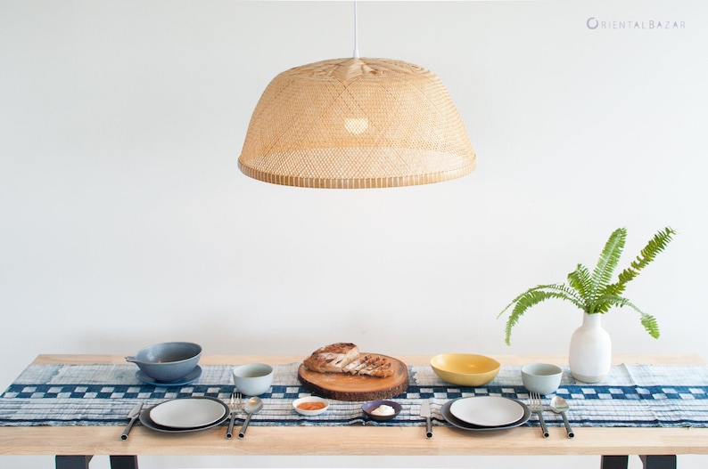 May include: A table setting with a woven bamboo pendant light hanging above. The table is set with two place settings, each with a white plate, a gray bowl, and silverware. A wooden cutting board with a slice of bread sits in the center of the table. A blue and white patterned table runner covers the table.