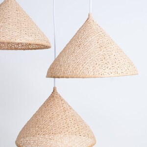 May include: Three woven, cone-shaped pendant lights hang from white cords against a white background. The lights are made of natural, light brown material and have a delicate, intricate pattern.
